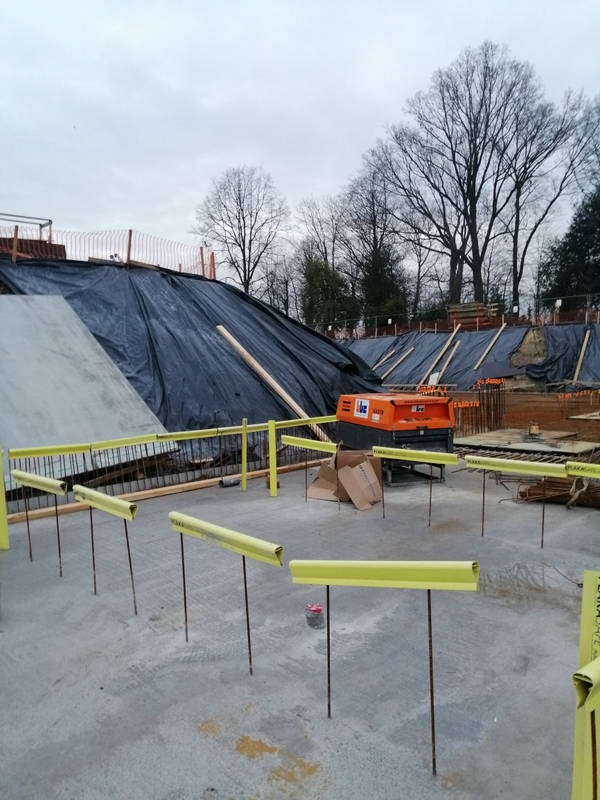 Betonowa płyta z żółtymi barierkami, w tle zabezpieczone folią zbocze i sprzęt budowlany. Widok na plac budowy w pochmurny dzień.