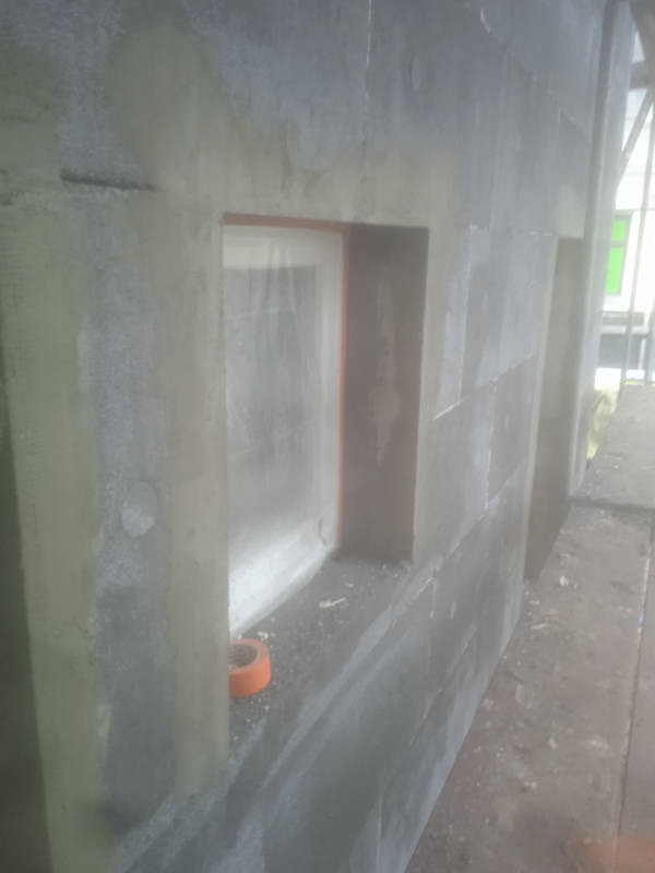 Ocieplona ściana budynku z widocznym oknem zabezpieczonym folią. Fragment pomarańczowej taśmy leży na parapecie. Widok z boku, z bliska.