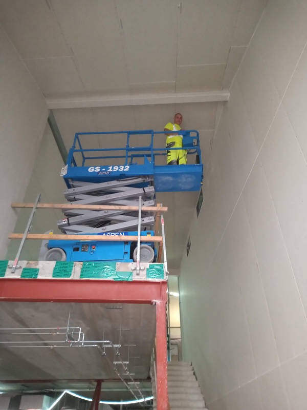 Pracownik na podnośniku GS-1932 montuje płyty karton gips na wysokiej ścianie wewnątrz budynku. Widoczne rusztowanie i schody. Oświetlenie sufitowe.