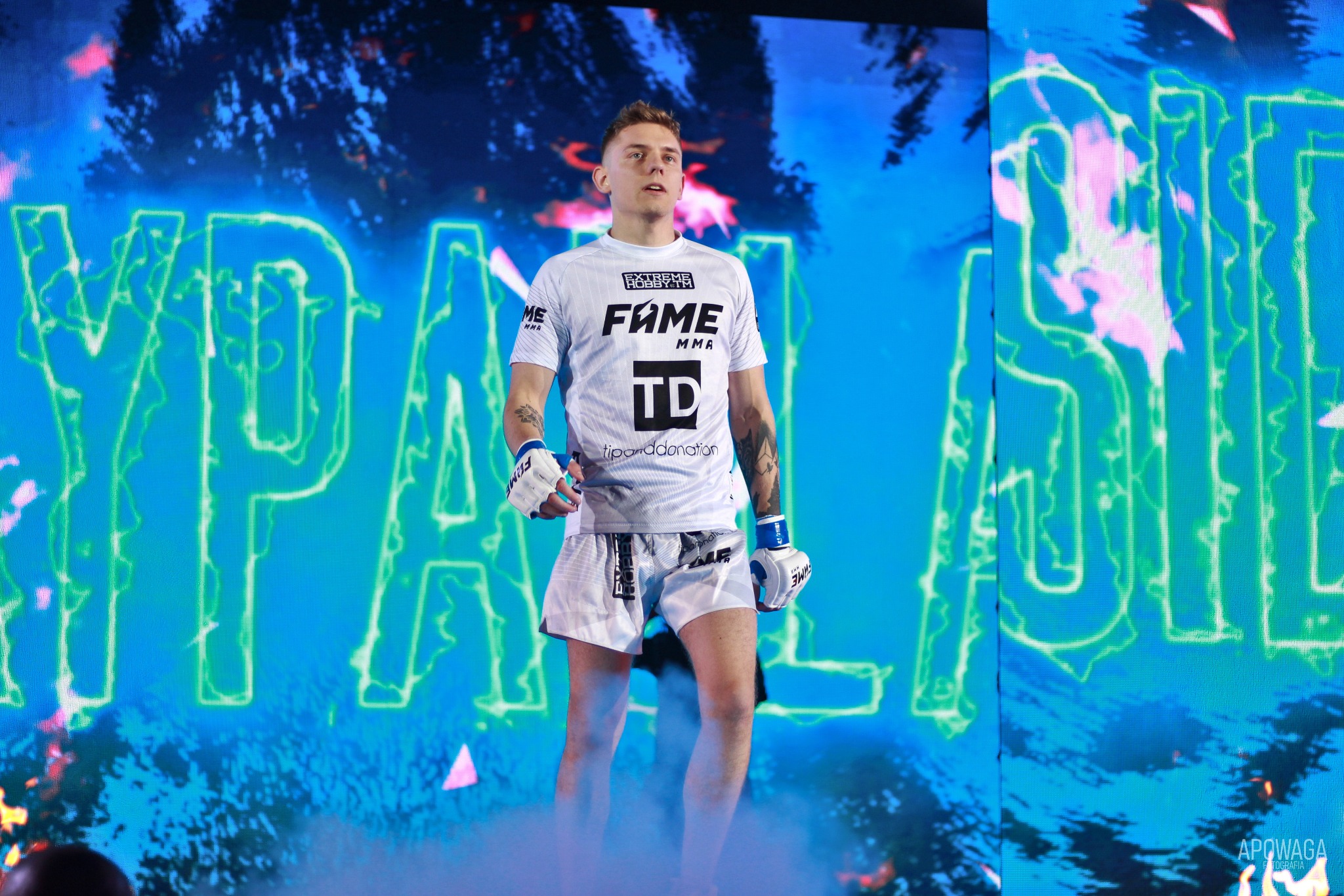 Młody zawodnik MMA w białym stroju z logo FAME MMA, wchodzi na arenę na tle podświetlonego ekranu LED z zielonymi napisami i efektem dymu.