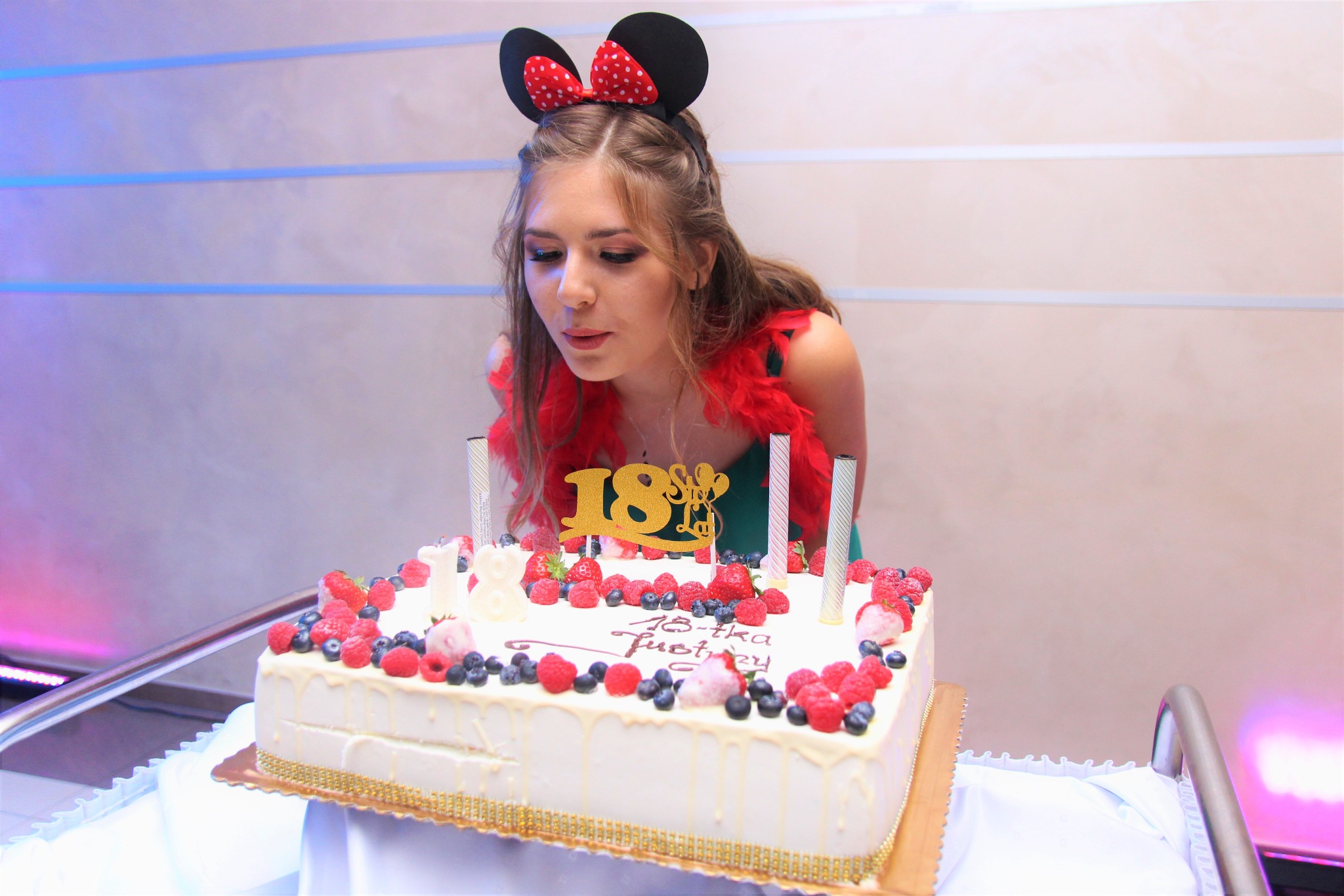 Młoda kobieta w opasce z uszami Myszki Miki dmucha świeczki na prostokątnym torcie urodzinowym udekorowanym owocami i napisem '18-tka Justyny'.