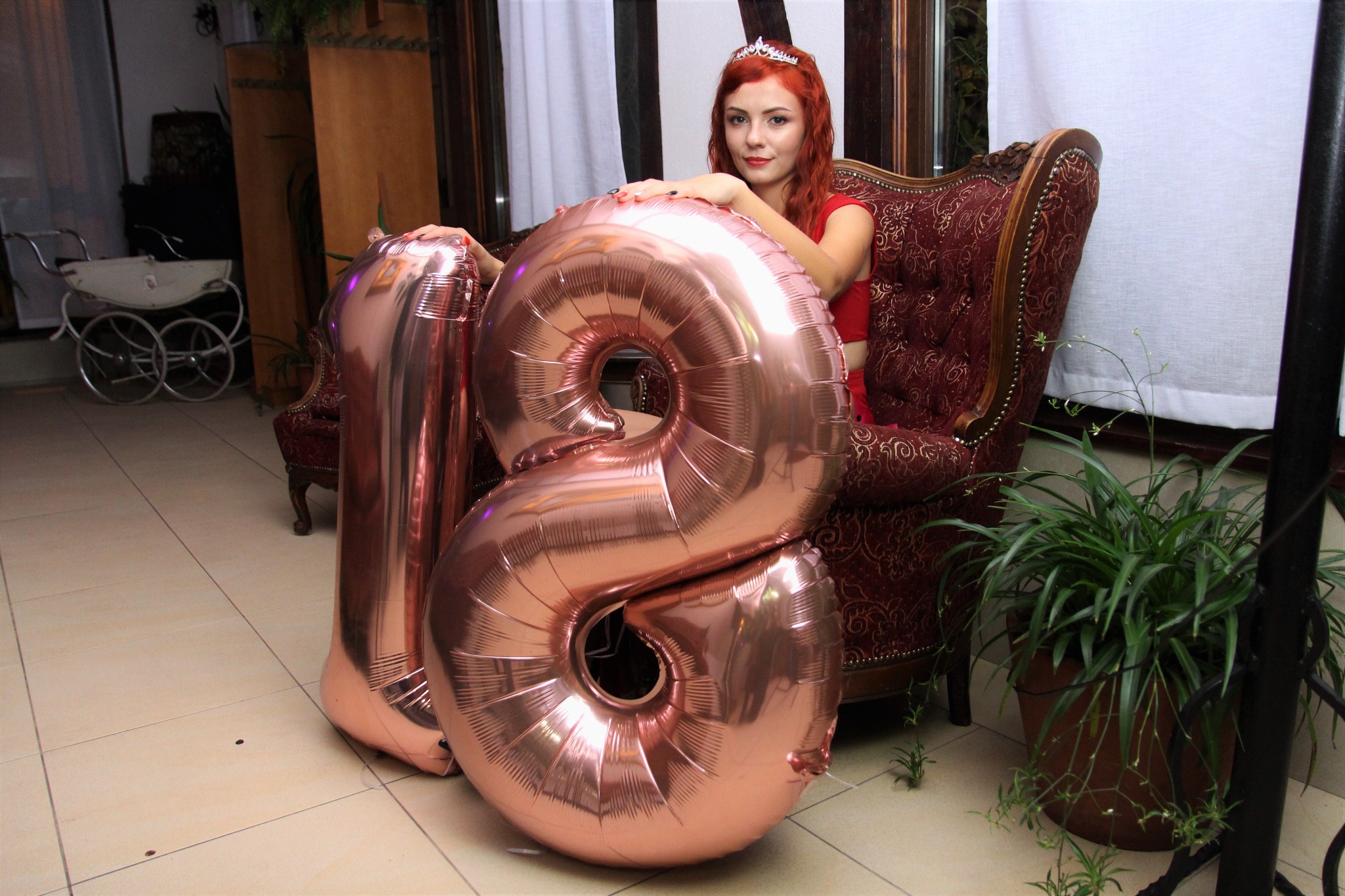 Uśmiechnięta, ruda dziewczyna w czerwonej sukience i diademie siedzi na bogato zdobionym, bordowym fotelu, opierając ręce na dużych, różowych balonach w kształcie cyfr '18'. W tle widać fragment...