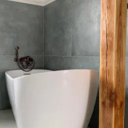 Nowoczesna łazienka z wolnostojącą białą wanną, szarą płytką imitującą beton i drewnianym słupem z widocznymi słojami, armatura w kolorze miedzi.