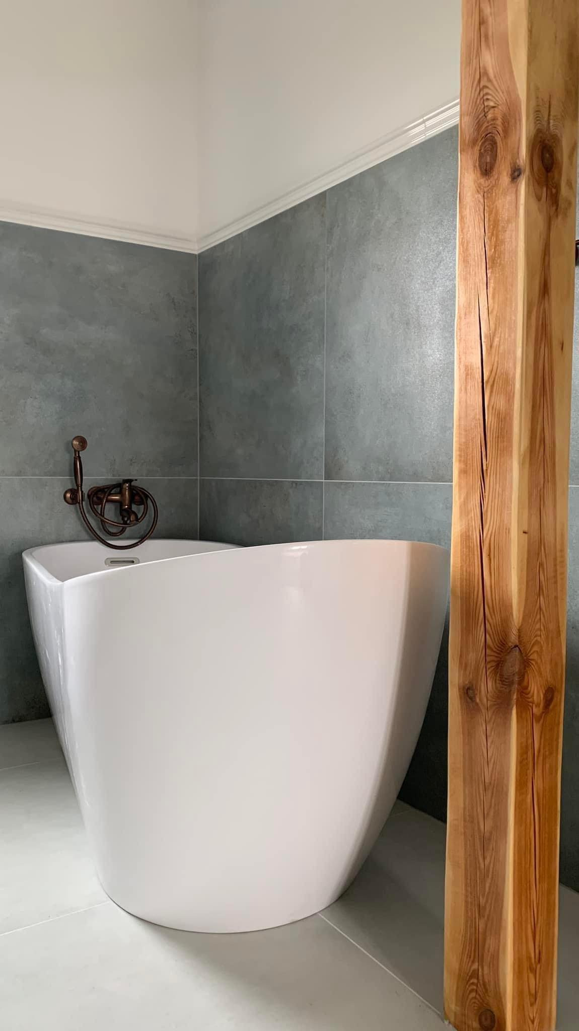 Nowoczesna łazienka z wolnostojącą białą wanną, szarą płytką imitującą beton i drewnianym słupem z widocznymi słojami, armatura w kolorze miedzi.