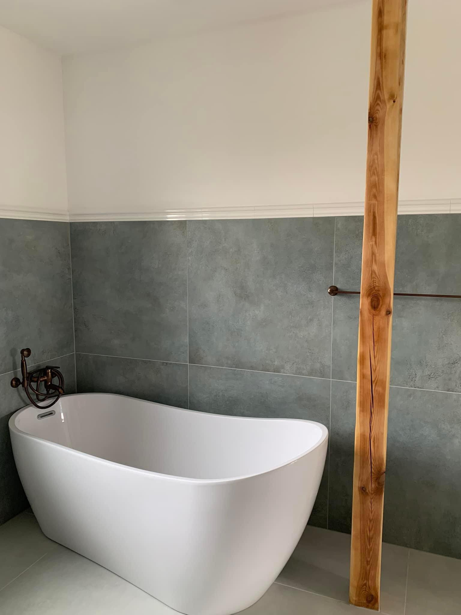 Nowoczesna łazienka z wolnostojącą białą wanną, szarymi płytkami imitującymi kamień, ozdobną armaturą w stylu retro i pionową drewnianą belką dekoracyjną z widocznymi słojami.