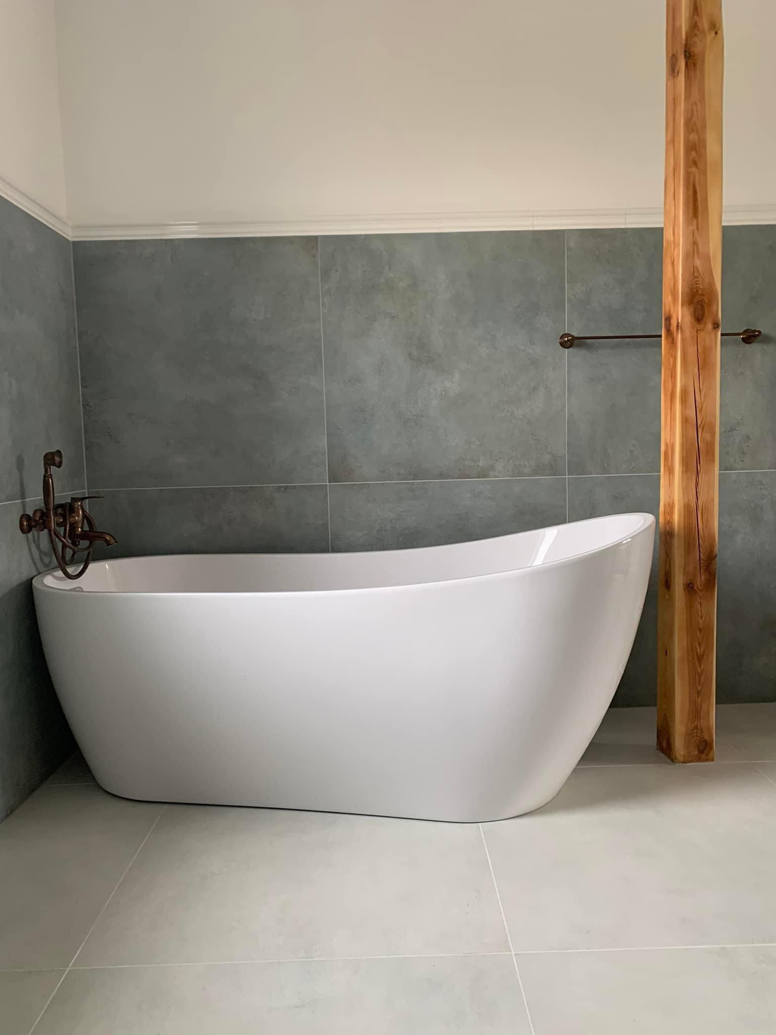 Wolnostojąca wanna w łazience wyłożonej szarymi, kwadratowymi płytkami. Mosiężna bateria wannowa w stylu retro, drewniany filar i minimalistyczny wieszak na ręczniki.