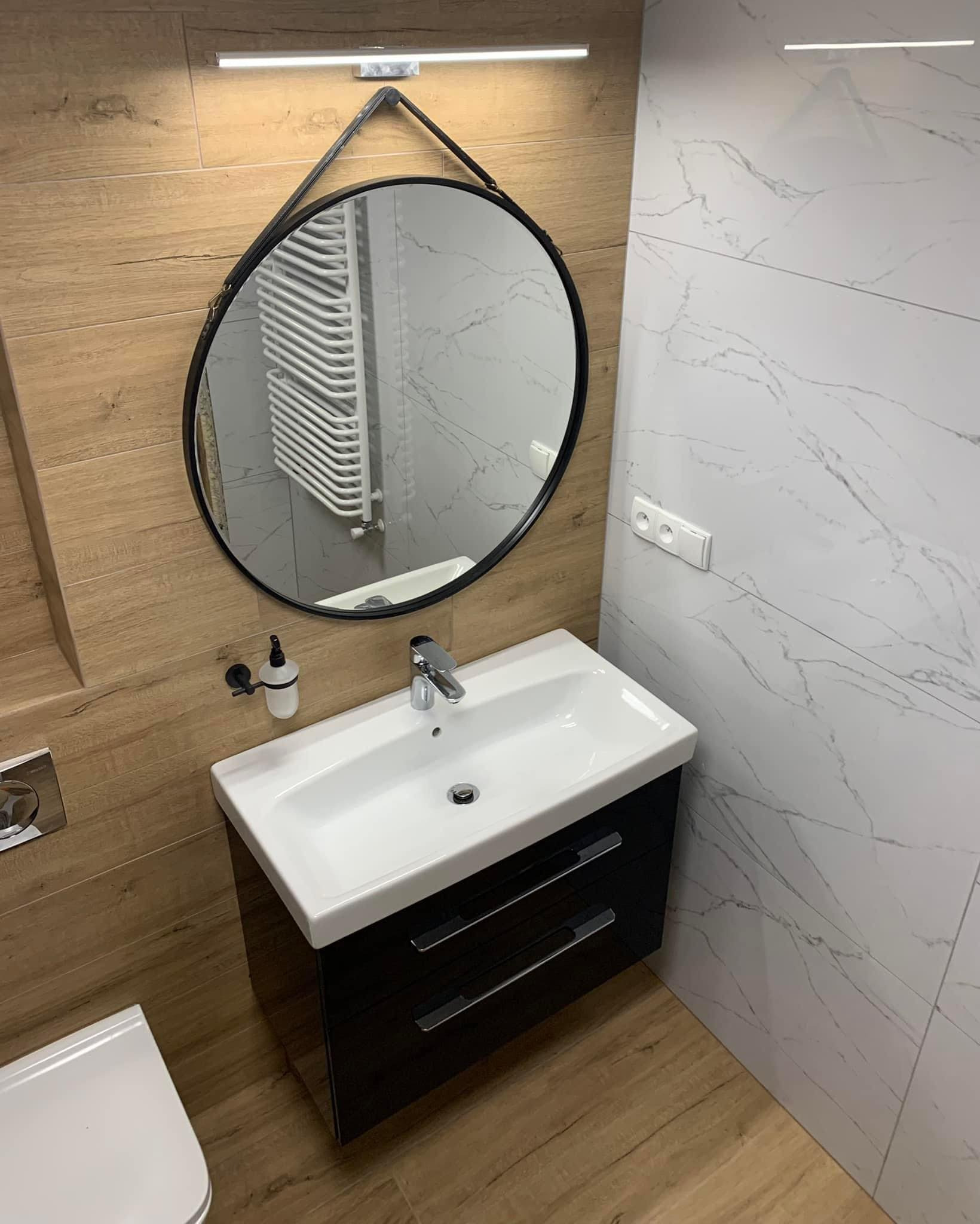 Nowoczesna łazienka z drewnopodobnymi i marmurowymi płytkami, okrągłym lustrem z czarną ramą i białą umywalką z szafką, oświetlona listwą LED