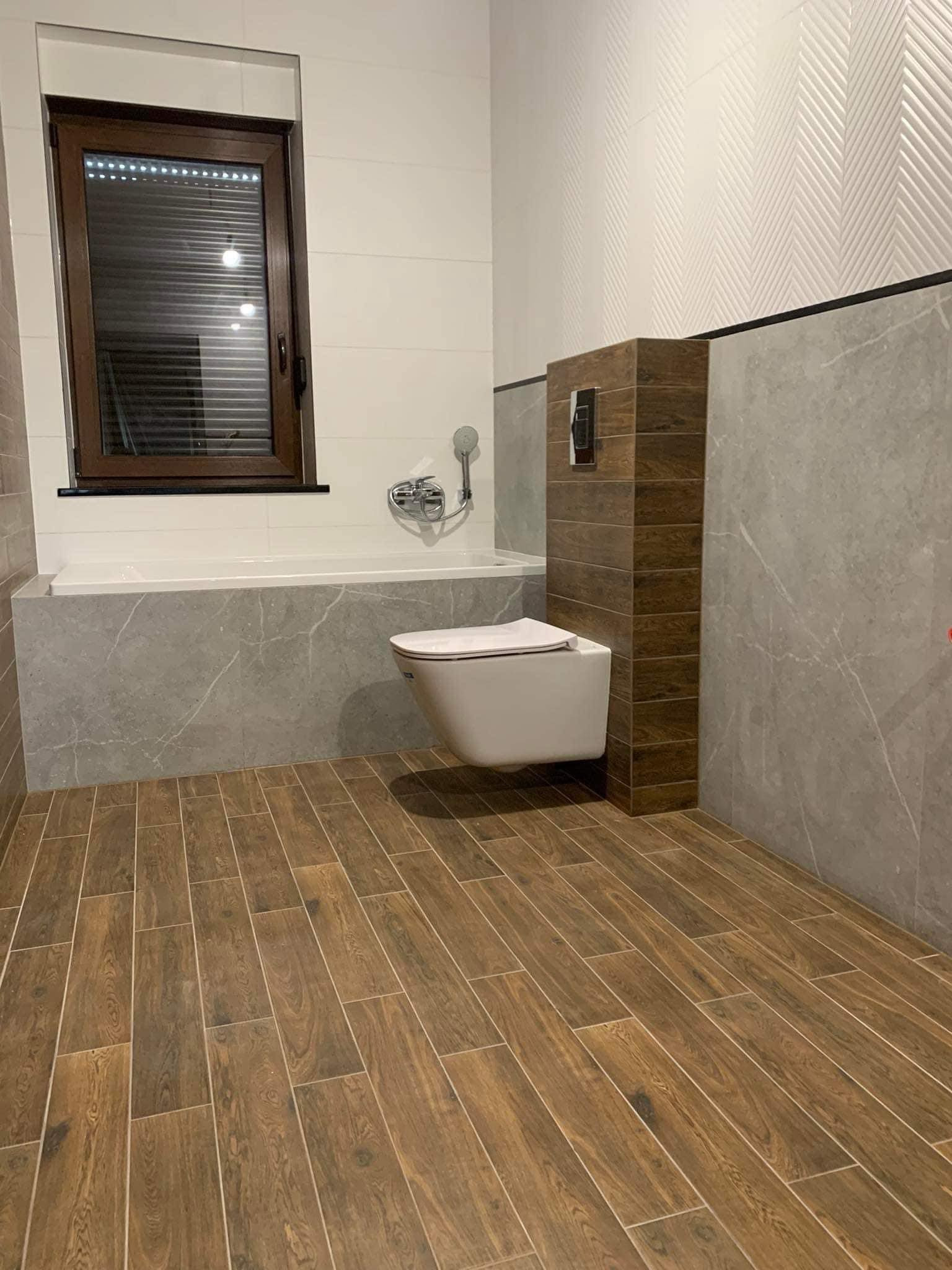 Wykończona łazienka z drewnopodobnymi płytkami podłogowymi, wiszącą toaletą z zabudową z płytek imitujących drewno, wanną obudowaną płytkami imitującymi marmur i ścianami wykończonymi białymi...