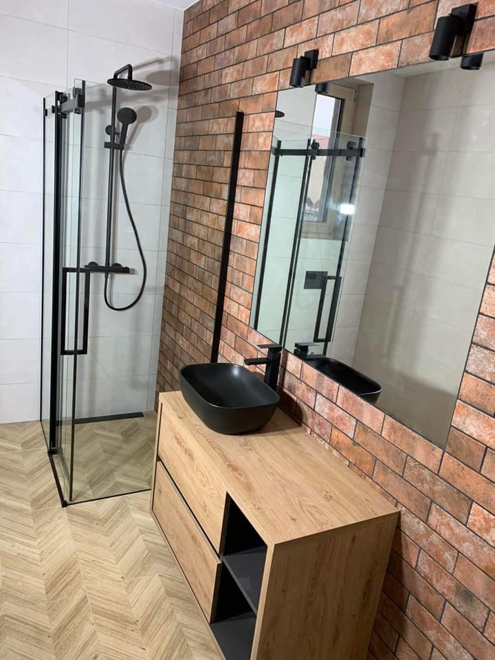 Nowoczesna łazienka z kabiną prysznicową typu walk-in z czarnymi profilami, umywalką nablatową w kolorze czarnym na drewnopodobnej szafce i ścianą wyłożoną cegłą.