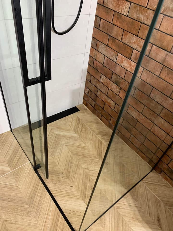 Nowoczesna kabina prysznicowa z czarnymi profilami, jasną podłogą ułożoną we wzór jodełki i ścianą wyłożoną płytkami imitującymi czerwoną cegłę.