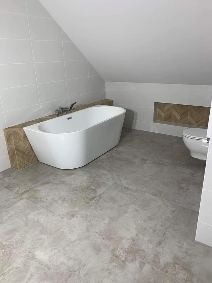 Wykończona łazienka na poddaszu z wolnostojącą wanną, szarymi płytkami podłogowymi imitującymi kamień, białymi płytkami ściennymi i wnęką z dekoracyjnym wzorem jodełki.