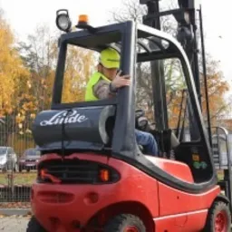 Operator w żółtym kasku i kamizelce odblaskowej prowadzi czerwony wózek widłowy Linde na placu manewrowym. Na widłach paleta, a przed wózkiem pomarańczowy pachołek. W tle budynki i drzewa.