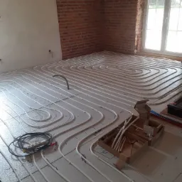 Rozłożone rury ogrzewania podłogowego na izolacji, przed wylaniem posadzki, widoczne złącza, narzędzia i elementy montażowe w kartonach, ściana z cegły w tle.