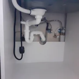 Syfon zlewozmywakowy z białego PCV, wężyki doprowadzające wodę i zawory odcinające zamontowane w szafce pod zlewem.