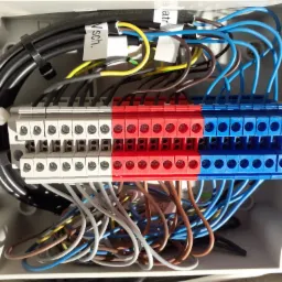Rozdzielnica elektryczna z przewodami oznaczonymi etykietami, widoczne listwy zaciskowe w kolorach białym, czerwonym i niebieskim, czarne opaski zaciskowe.