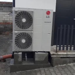 Zewnętrzna jednostka pompy ciepła LG Therma V, zamontowana na betonowym fundamencie przy ścianie budynku z szarą stolarką.