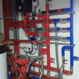 Rozbudowana instalacja centralnego ogrzewania z rurami w izolacji czerwonej, niebieskiej i szarej, z widocznym kotłem i osprzętem, zamontowana na białej ścianie.