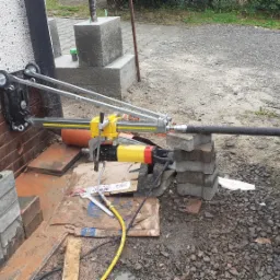 Sprzęt do wiercenia otworów w ścianie, z żółto-czarną wiertnicą rdzeniową, zamocowany do ściany za pomocą metalowej ramy z kółkami, ustawiony na cegłach, przewód przyłączeniowy do wiertnicy, obok...