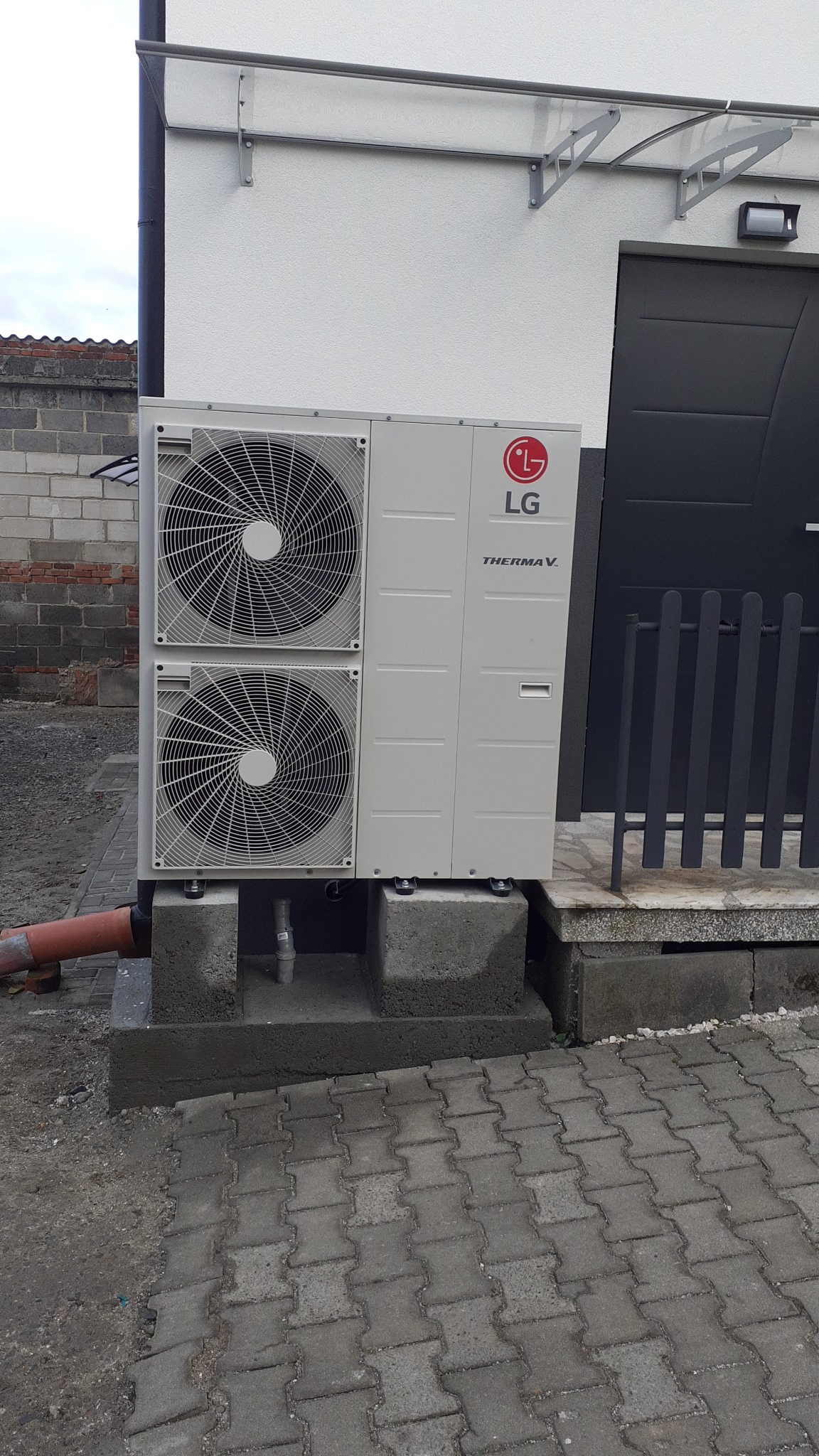 Zewnętrzna jednostka pompy ciepła LG Therma V, zamontowana na betonowym fundamencie przy ścianie budynku z szarą stolarką.