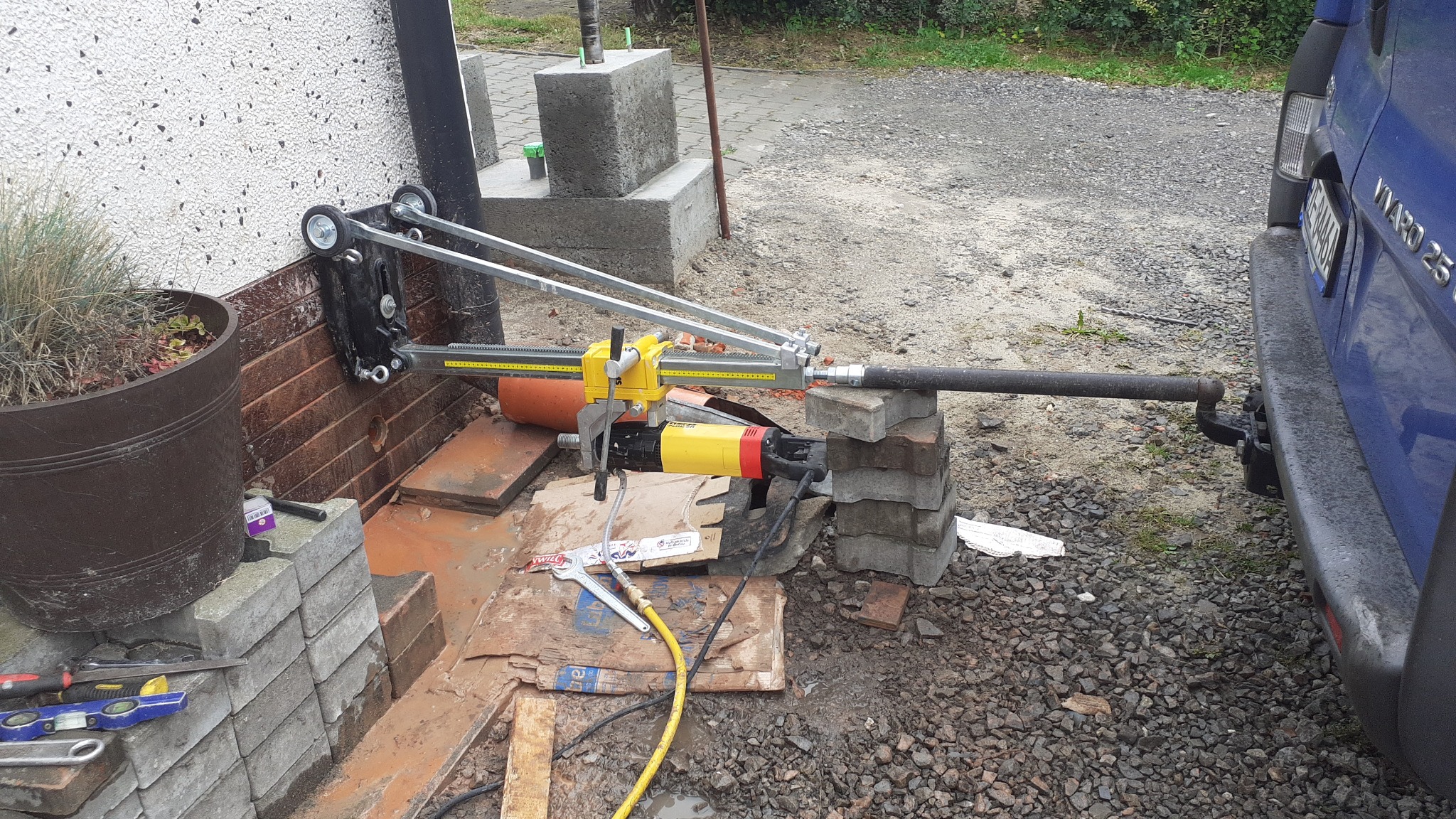 Sprzęt do wiercenia otworów w ścianie, z żółto-czarną wiertnicą rdzeniową, zamocowany do ściany za pomocą metalowej ramy z kółkami, ustawiony na cegłach, przewód przyłączeniowy do wiertnicy, obok...