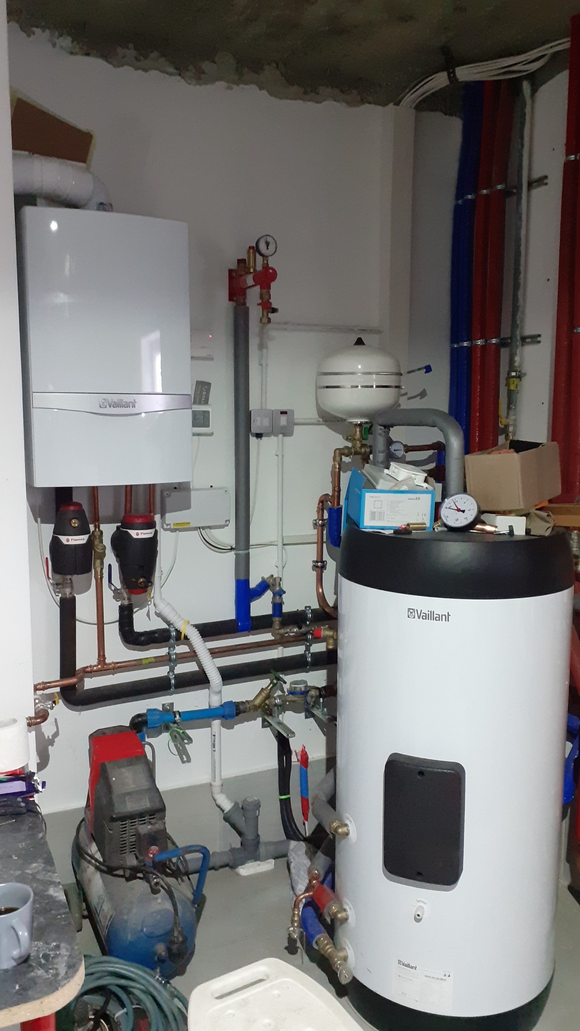 Biały piec CO Vaillant z podłączonymi rurami miedzianymi i plastikowymi, obok zbiornik ciśnieniowy, widoczny manometr, instalacja w surowym stanie.