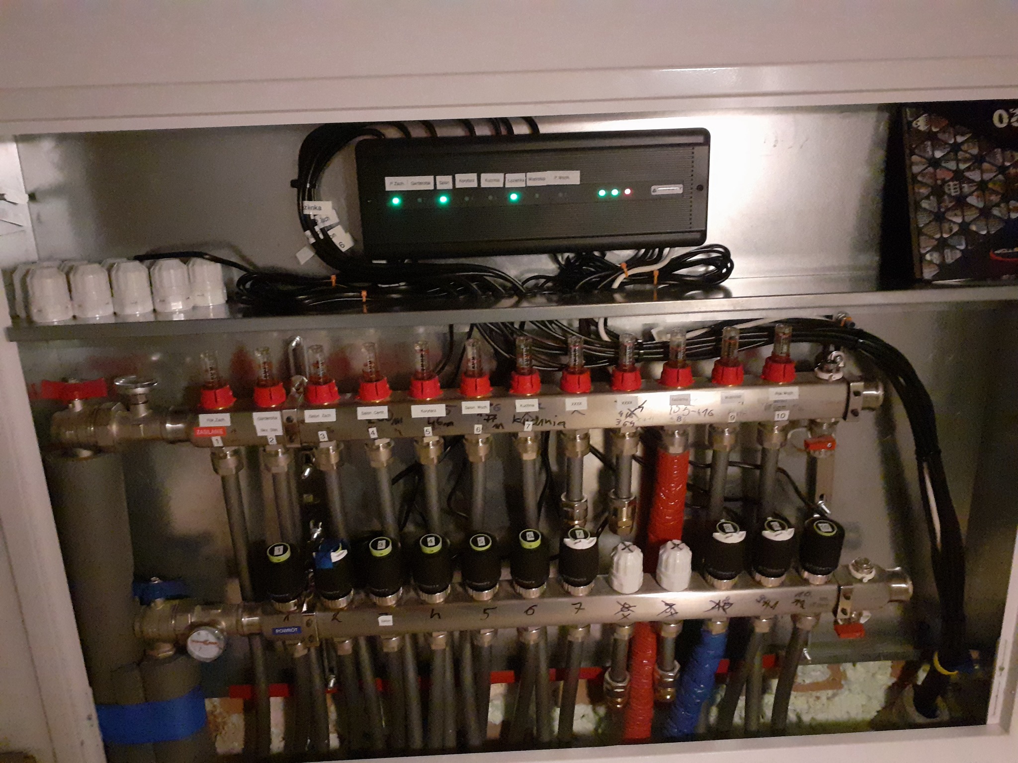 Rozdzielacz centralnego ogrzewania w szafce, z elektronicznym sterownikiem i przewodami, widoczne zawory z opisami stref grzewczych.