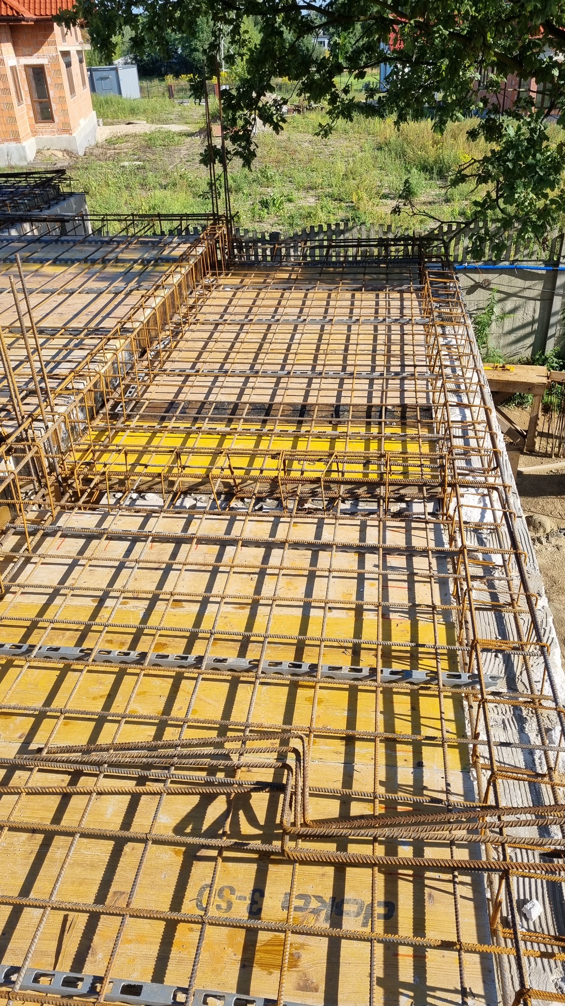 Siatka zbrojeniowa ułożona na żółtym szalunku, przygotowana do zalania betonem, widok z góry, w tle budynek w stanie surowym otwartym i zieleń.