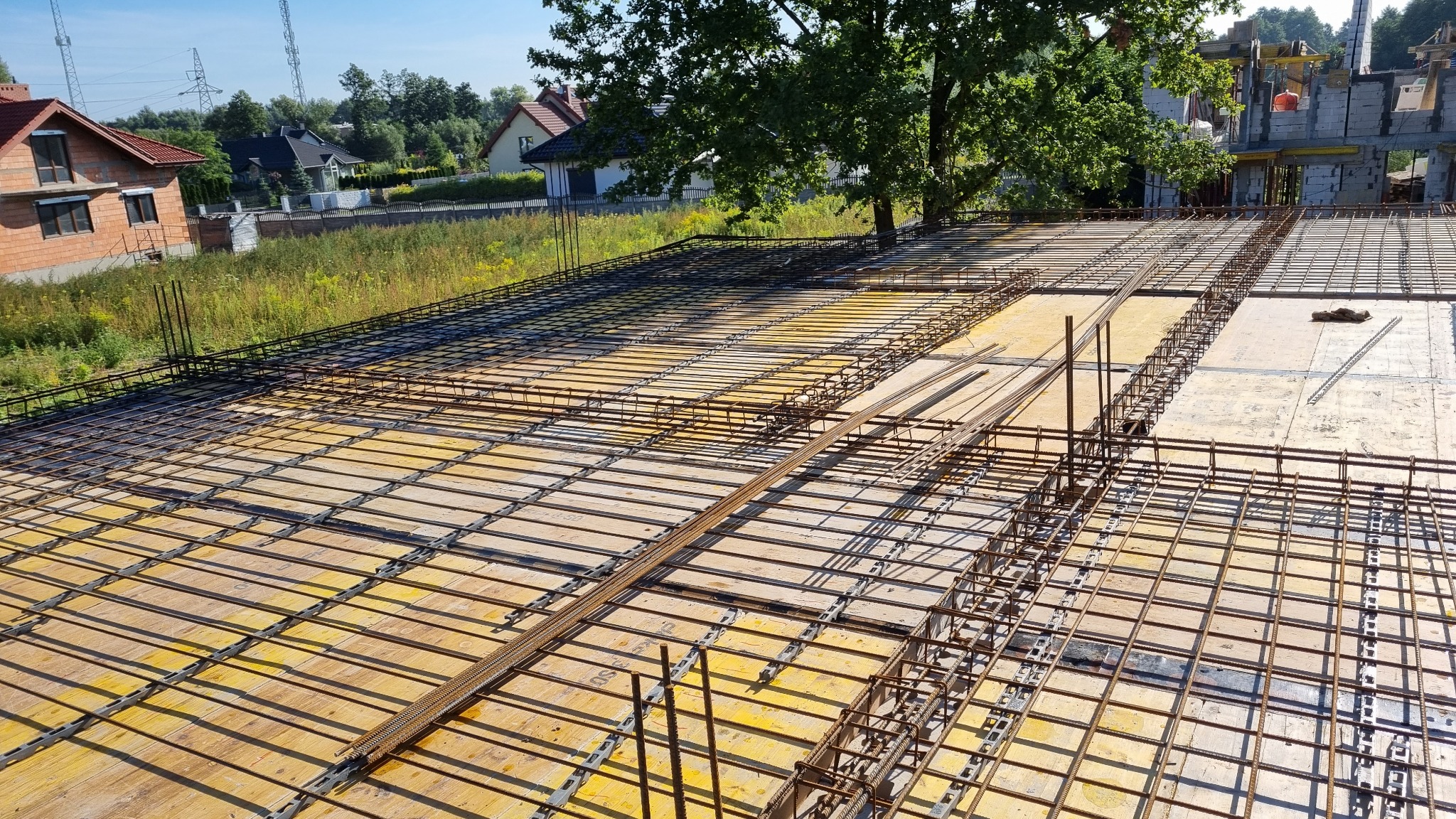 Zbrojenie stropu na żółtym deskowaniu z widocznymi prętami i siatkami stalowymi, w tle budynek w trakcie budowy i zieleń.