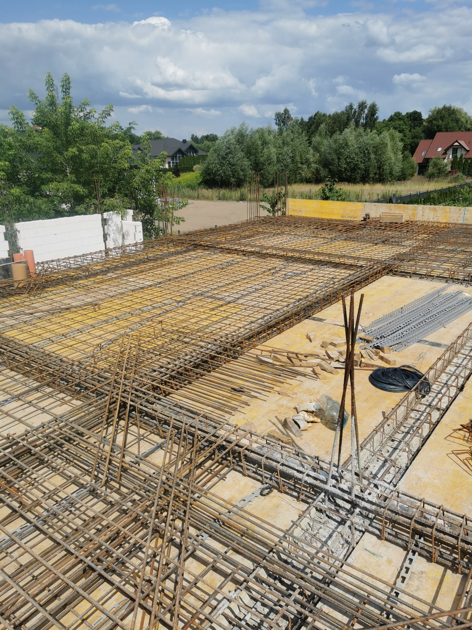 Siatka zbrojeniowa ułożona na żółtym szalunku, przygotowana do zalania betonem, widok z góry na plac budowy z fragmentami zieleni i domów w tle.