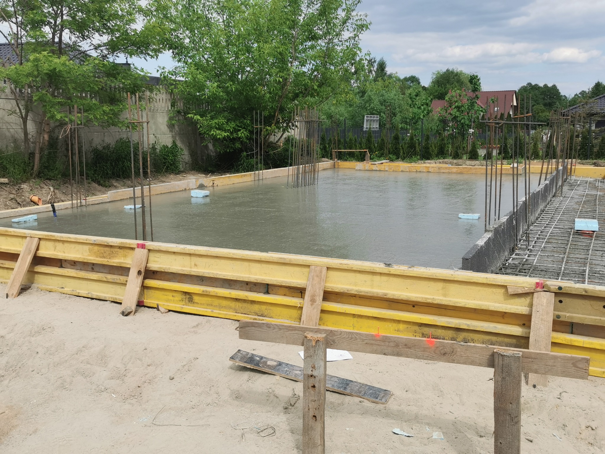 Wylany fundament z żółtym szalunkiem i wystającymi prętami zbrojeniowymi, częściowo zalany wodą, z widocznymi elementami izolacji termicznej i drewnianymi podporami na piaszczystym podłożu.