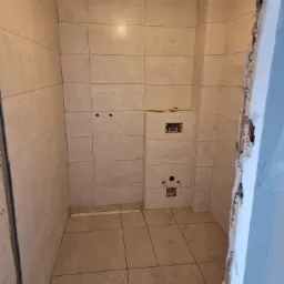 Wnętrze łazienki w trakcie remontu, widoczne ułożone płytki na ścianach i podłodze, przygotowane otwory na instalacje sanitarne i elektryczne, surowy otwór drzwiowy.