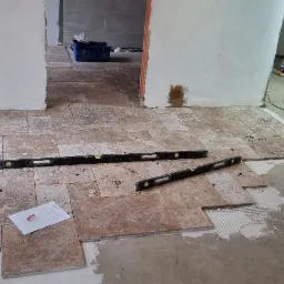 Układanie trawertynu na podłodze w trakcie remontu, widoczne poziomice, klej i fragment planu pomieszczenia.