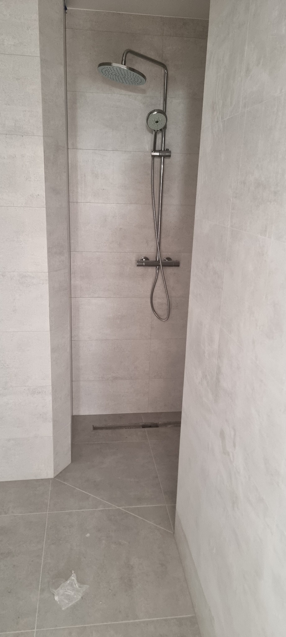 Nowoczesna kabina prysznicowa walk-in z szarymi płytkami, deszczownicą i odpływem liniowym w podłodze, widoczne niedociągnięcia na podłodze.