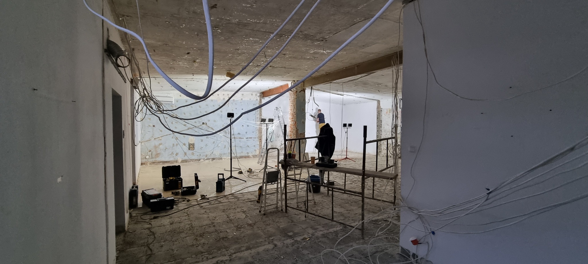 Wnętrze w trakcie remontu: widoczne kable zwisające z sufitu, rusztowanie z pracownikiem, sprzęt oświetleniowy i narzędzia, ściany w stanie surowym.