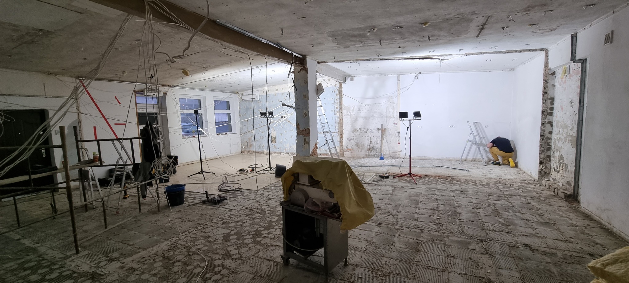 Wnętrze w trakcie gruntownego remontu: zerwane podłogi, odsłonięte ściany, wiszące kable, drabiny i reflektory budowlane, pracownik w żółtych butach przy ścianie.