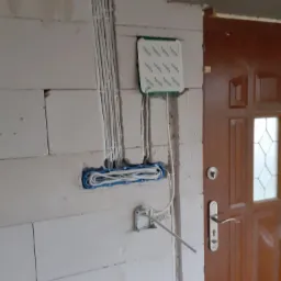 Ściana z betonu komórkowego w trakcie prac instalacyjnych: widoczne kable w peszlach, puszka elektryczna z zielonymi oznaczeniami i wystającymi przewodami oraz metalowa puszka pod gniazdko obok...