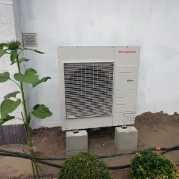 Biała pompa ciepła Immergas, zamontowana na betonowych bloczkach przy białej ścianie budynku, obok rosnący słonecznik w początkowej fazie wzrostu.