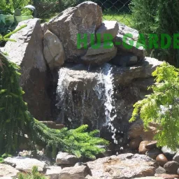 Ogród skalny z kaskadą wodną, otoczony zielenią. Woda spływa po kamieniach do małego stawu z otoczakami. Dekoracyjne iglaki dodają naturalnego uroku.