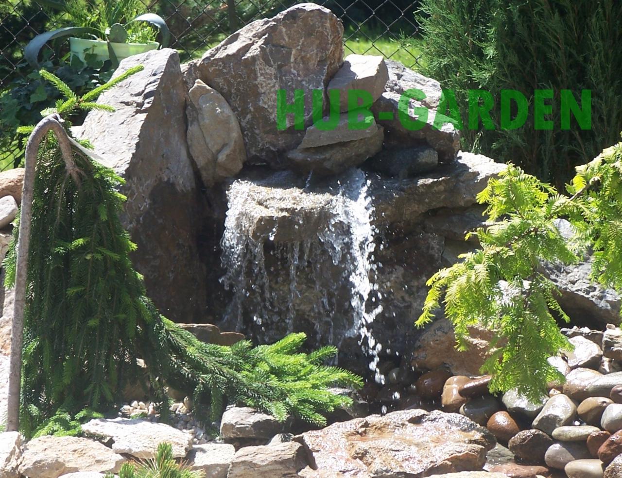 Ogród skalny z kaskadą wodną, otoczony zielenią. Woda spływa po kamieniach do małego stawu z otoczakami. Dekoracyjne iglaki dodają naturalnego uroku.