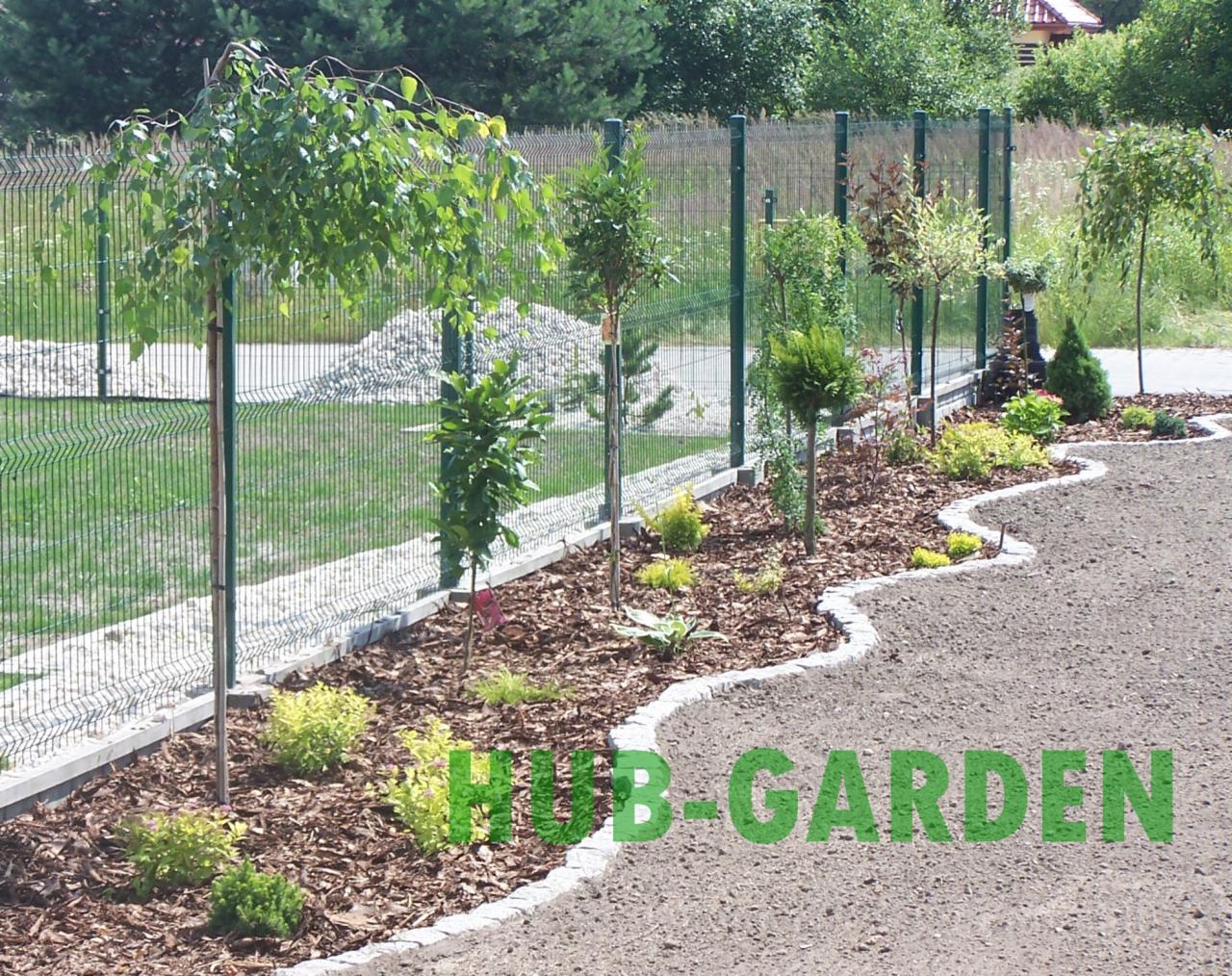 Aranżacja ogrodu z młodymi drzewami i krzewami, kora na rabatach, obrzeże z kostki, siatka ogrodzeniowa w tle. Widok na świeżo założony ogród.