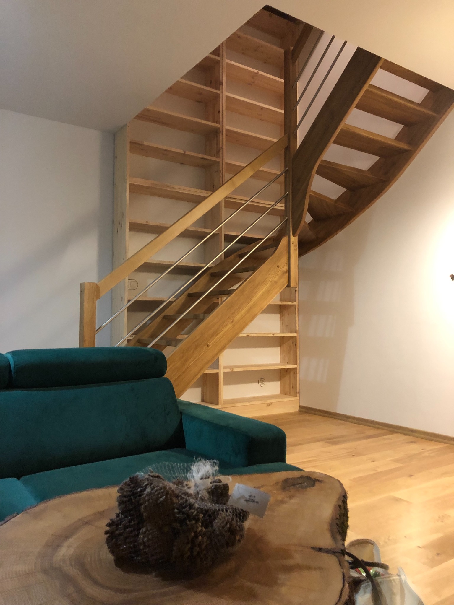 Drewniane schody z metalowymi barierkami integrujące się z regałem na książki, obok zielona sofa i stolik kawowy z drewna.