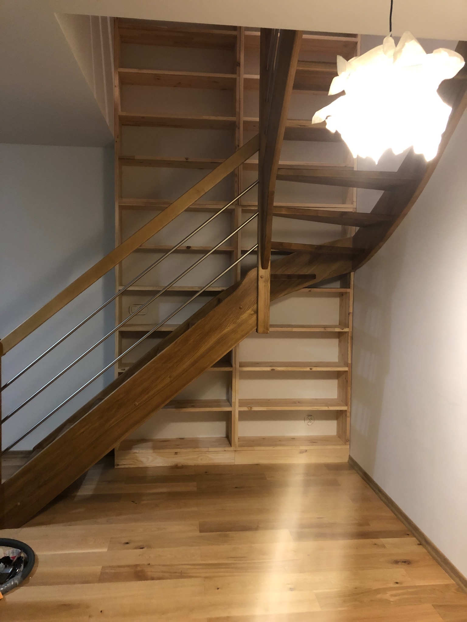 Drewniane schody z metalową balustradą wkomponowane w regał z jasnego drewna, oświetlone ozdobną lampą, parkiet na podłodze.