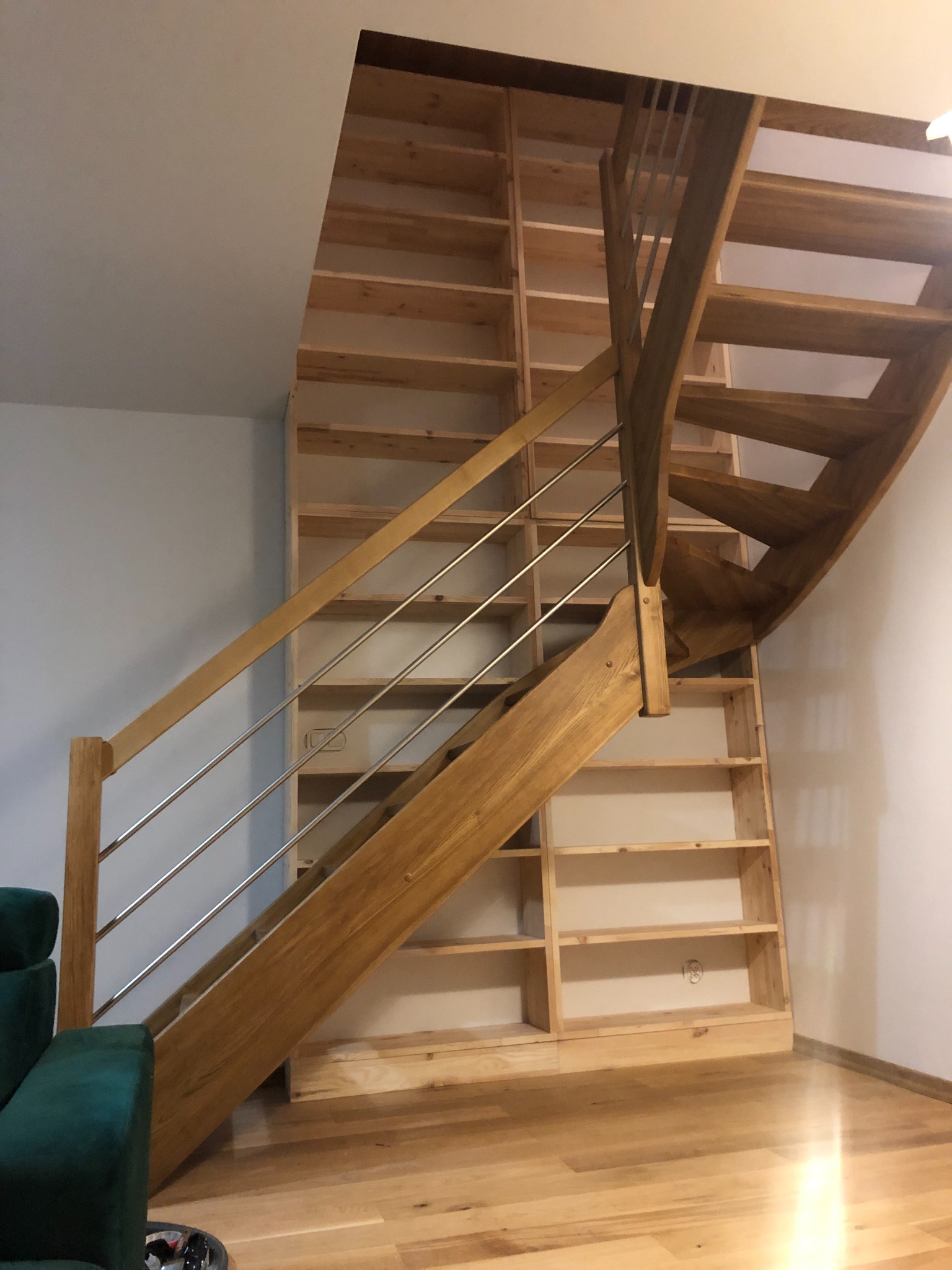 Drewniane schody z metalową balustradą prowadzące do drewnianego regału w zabudowie, widoczne z dołu, z fragmentem zielonego fotela w lewym dolnym rogu.