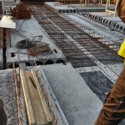 B.R.T- BP Sp. z o.o. - Konstrukcja stropu żelbetowego z ułożonym zbrojeniem i elementami szalunku, widok z perspektywy pracownika w odzieży roboczej, w tle drzewa i zachodzące słońce.