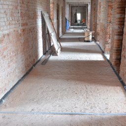 B.R.T- BP Sp. z o.o. - Wnętrze budynku w trakcie budowy z surowymi ceglanymi ścianami, piaskową podłogą, instalacją przewodów wzdłuż ścian i rolką kabla w oddali.