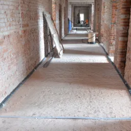 Wnętrze budynku w trakcie budowy z surowymi ceglanymi ścianami, piaskową podłogą, instalacją przewodów wzdłuż ścian i rolką kabla w oddali.