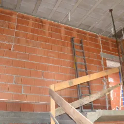 Wnętrze domu w trakcie budowy: ściany z czerwonej cegły, częściowo położone kable elektryczne, drabina oparta o ścianę, drewniane szalunki schod&oacute;w i podpory stropu.