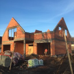 B.R.T- BP Sp. z o.o. - Wznoszenie dwukondygnacyjnego budynku z czerwonej cegły, widoczny szkielet konstrukcji, okna bez ram, pracownik wewnątrz, palety z materiałami budowlanymi i kontener na gruz przed budynkiem.