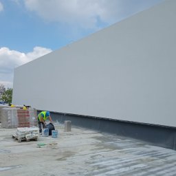 B.R.T- BP Sp. z o.o. - Pracownik w kamizelce odblaskowej miesza zaprawę na dachu budynku z ocieploną ścianą szczytową, widoczne paczki wełny mineralnej.