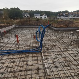 B.R.T- BP Sp. z o.o. - Zbrojenie płyty fundamentowej z wystającymi rurami instalacyjnymi w kolorze pomarańczowym i niebieskim, widok na budynki w tle.